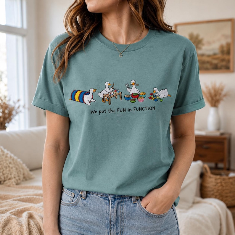 May include: A dusty blue t-shirt featuring a graphic of four white ducks engaged in various activities, including crawling through a tunnel, crafting at a table, eating, and playing with blocks, with the text "We put the FUN in FUNCTION" below.
