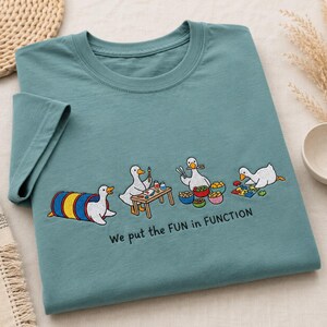 May include: A teal short-sleeved t-shirt featuring an embroidered graphic of four white ducks engaged in various activities, including playing in a tunnel, painting, eating, and doing a puzzle, with the text "We put the FUN in FUNCTION".