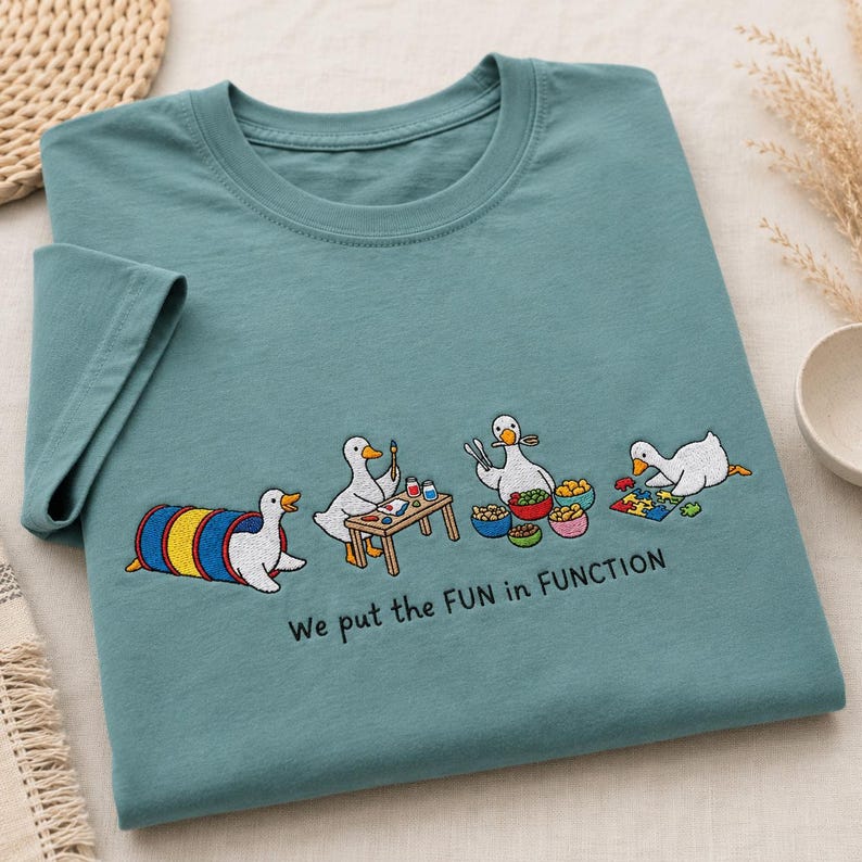 May include: A teal short-sleeved t-shirt featuring an embroidered graphic of four white ducks engaged in various activities, including playing in a tunnel, painting, eating, and doing a puzzle, with the text "We put the FUN in FUNCTION".