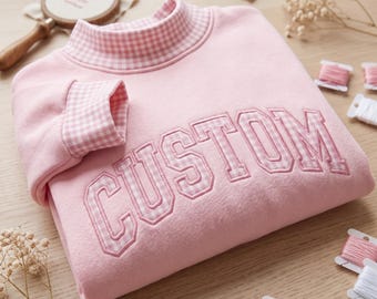 May include: A light pink sweatshirt featuring a mock neck collar and sleeve cuffs in a pink and white gingham pattern. The word CUSTOM is embroidered across the chest in a varsity-style font with a pink and white gingham fabric fill.