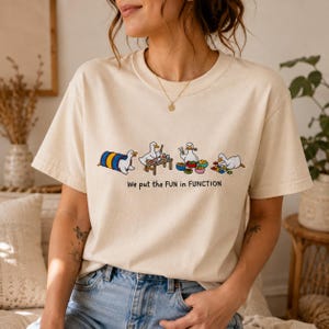 May include: A cream-colored short-sleeve t-shirt featuring a graphic of four white ducks engaged in various activities. Below the ducks, the text reads, "We put the FUN in FUNCTION." The shirt has a crew neckline and a relaxed, casual fit.