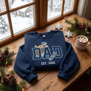Könnte beinhalten: Ein marineblaues Sweatshirt mit Rundhalsausschnitt und einer Stickerei, die eine große blaue Schildkröte und eine kleine blaue Schildkröte zeigt. Der Schriftzug DAD ist in großen hellblauen Buchstaben mit EST. 2026 darunter abgebildet.