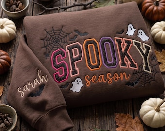 May include: A brown sweatshirt featuring embroidered text that reads SPOOKY season in colorful block and script lettering. The design includes embroidered white ghosts, black spiderwebs, and bats. The sleeve is embroidered with the name Sarah.