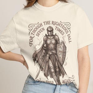 以下が含まれることがあります： オフホワイトの半袖Tシャツ。剣と盾を持った鎧姿の騎士のダークブラウンのグラフィックが描かれています。騎士の周りには「FOR THOUGH THE RIGHTEOUS FALL SEVEN TIMES THEY RISE AGAIN.」というテキストが記されています。