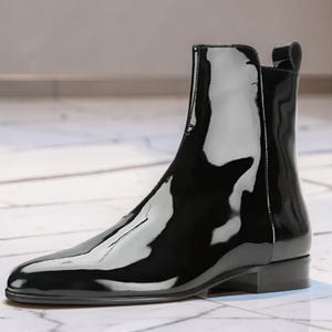 May include: A black ankle boot with a high-gloss patent finish, featuring a sleek silhouette, a pointed toe, a low stacked heel, and a pull tab at the back of the collar. The boot has a smooth, reflective surface and a clean, minimalist design.