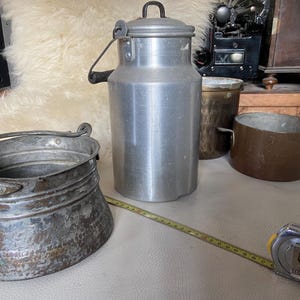 Pode incluir: Uma coleção de utensílios de cozinha vintage, incluindo um balde de metal desgastado com alça, uma lata de leite alta de alumínio prateado com tampa e duas panelas cor de cobre. Uma fita métrica amarela está estendida para mostrar a escala.