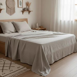 May include: A light beige linen bed sheet set featuring a flat sheet draped over a mattress and two matching pillowcases on a wooden bed frame. The fabric has a visible woven texture and a neutral, solid color tone throughout the bedding set.