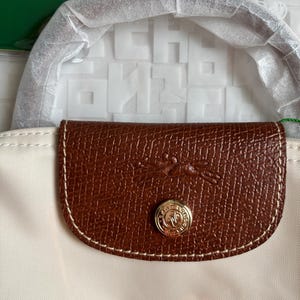 May include: A cream-colored handbag featuring a brown leather flap with a debossed horse and rider logo. The flap includes a gold-tone metal snap button with engraved text. The handle is wrapped in protective white paper, and the bag is partially covered.