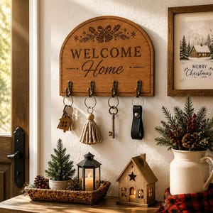 Könnte beinhalten: Ein halbkreisförmiger, hölzerner Wand-Schlüsselhalter mit der Aufschrift "WELCOME Home" und einer Glockengravur. Er verfügt über vier Haken aus antikem Metall, an denen jeweils ein Schlüsselbund hängt, inklusive eines Hausanhängers.