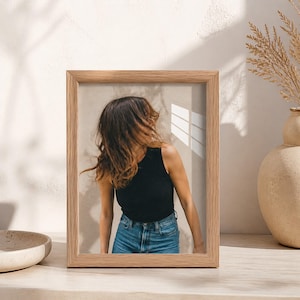 Könnte beinhalten: Ein rechteckiger Bilderrahmen aus hellem Holz mit einem Foto einer Person mit schulterlangem braunem Haar, die ein schwarzes ärmelloses Oberteil und eine blaue Jeans trägt, platziert vor einer neutralen Wand mit weichen Lichtschatten.