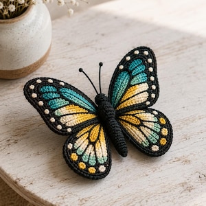 May include: A handcrafted crochet butterfly featuring a black body and antennae, with wings displaying a vibrant gradient of teal, yellow, and cream colors, accented by small white and yellow dots along the black outer edges of the wings.