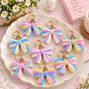 May include: A collection of eight handmade crochet bow keychains in various pastel rainbow colour combinations. Each bow features a gold-tone metal keyring, a small heart charm, and a single round bead in colours like pink, blue, or yellow.