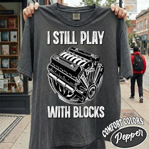 May include: A charcoal grey short-sleeve t-shirt featuring a white graphic illustration of a V8 engine block in the centre. The text "I STILL PLAY" is printed above the engine graphic, and the text "WITH BLOCKS" is printed below the graphic.
