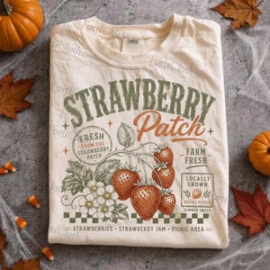 以下が含まれることがあります： Strawberry Patchという文字がプリントされたクリーム色のTシャツ。イチゴ、イチゴの花、葉のイラストが描かれており、Fresh From The Strawberry PatchやFarm Freshという文字も含まれています。