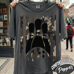 May include: A charcoal gray short-sleeve t-shirt featuring a central graphic of three black bowling pins arranged in a row. The graphic is overlaid on a vertical camouflage pattern in shades of brown, tan, and black, resembling a flag design.