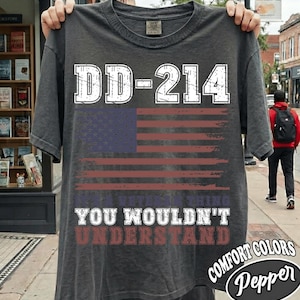 May include: A charcoal gray short-sleeve t-shirt featuring a distressed graphic of the American flag. The text DD-214 is printed in large white letters above the flag, with the text IT'S A VETERAN THING YOU WOULDN'T UNDERSTAND below it.