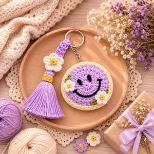 Op de afbeelding: Een ronde, lavendelkleurige gehaakte sleutelhanger met een zwart geborduurd smileygezicht en twee kleine witte bloemen. De sleutelhanger bevat een gevlochten lavendelkleurig koord, een geel omwikkelde kwast en een zilveren metalen ring.