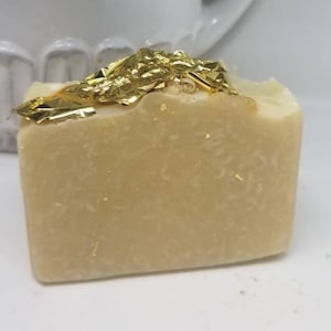 Puede incluir: Una barra de jabón rectangular con copos de oro en la parte superior. El jabón es de color beige claro.