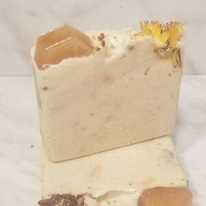 Puede incluir: Dos barras de jabón hechas a mano con un diseño floral. La barra superior tiene una flor seca y un trozo de jabón naranja en la parte superior. La barra inferior tiene una flor seca y un trozo de jabón naranja en el lateral.