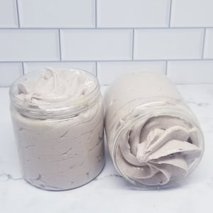 Könnte beinhalten: Zwei transparente Gläser gefüllt mit helllila, geschlagenem Körperbutter. Die Gläser sind offen und zeigen das strukturierte, cremige Produkt. Der Hintergrund ist eine weiße geflieste Wand.