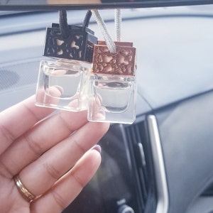 May include: Two small glass bottles with decorative metal tops hanging from a rearview mirror. The bottles are filled with a clear liquid and have a black and gold design.