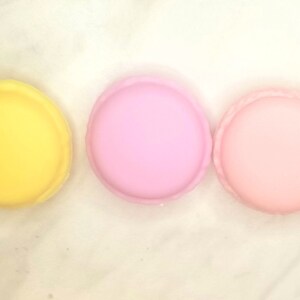 Puede incluir: Tres macarons redondos, uno amarillo, uno rosa claro y uno rosa oscuro, dispuestos en fila sobre una superficie blanca.