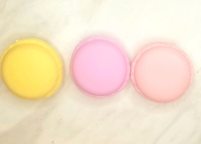 Puede incluir: Tres macarons redondos, uno amarillo, uno rosa claro y uno rosa oscuro, dispuestos en fila sobre una superficie blanca.