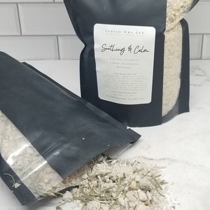 May include: A black resealable pouch filled with white bath salts and green herbs. The pouch has a label that reads "Indigo Owl LLC. Botanical Bath Salts. Soothing & Calm. Tea Tree, Turmeric, Lemon, Eucalyptus, Rosemary. Ingredients: Epsom Salt, Small Crystal Pink Himalayan Salt, Essential Oil Blend (Tea Tree, Turmeric, Lemon, Eucalyptus, Rosemary), Dried Lavender Buds, Dried Organic Calendula Flowers, Fine Sea Salt, Colloidal Oatmeal, Dried Rose Petals, Dried Tapioca Starch, Baking Soda. Net Wt. 14 oz. Handcrafted in the USA."