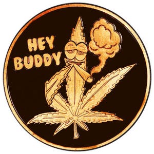 Op de afbeelding: Een ronde koperkleurige metalen munt met een gegraveerde afbeelding van een cannabisblad met een cartoongezicht dat een joint rookt, met een rookwolk erboven. De tekst HEY BUDDY is in een vetgedrukt lettertype aan de linkerkant gegraveerd.