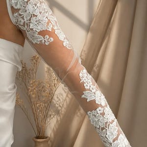 Peut inclure: Une longue manche blanche transparente ornée de broderies florales en dentelle complexe. Le motif délicat s'étend de l'épaule jusqu'au poignet, avec une conception ajustée qui recouvre le bras d'un tissu en maille semi-transparente.