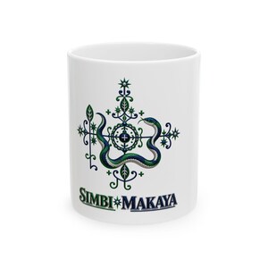May include: A white ceramic mug featuring a central graphic of a blue and green snake coiled around a symmetrical, ornate cross-like symbol with star accents. Below the graphic, the text SIMBI MAKAYA is printed in a bold, dark font.