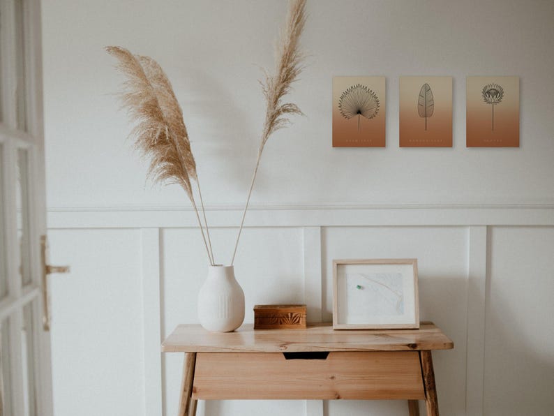 May include: Three rectangular wall art prints featuring black botanical line drawings of a palm leaf, a banana leaf, and a protea flower against a gradient background transitioning from cream to terracotta orange.