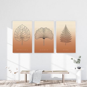 May include: A set of three rectangular wall art prints featuring black line art illustrations of a monstera leaf, a ginkgo leaf, and a fern leaf. Each print has a gradient background transitioning from light cream to a warm terracotta orange.