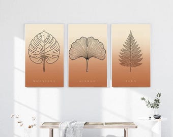 May include: A set of three rectangular wall art prints featuring black line art illustrations of a monstera leaf, a ginkgo leaf, and a fern leaf. Each print has a gradient background transitioning from light cream to a warm terracotta orange.