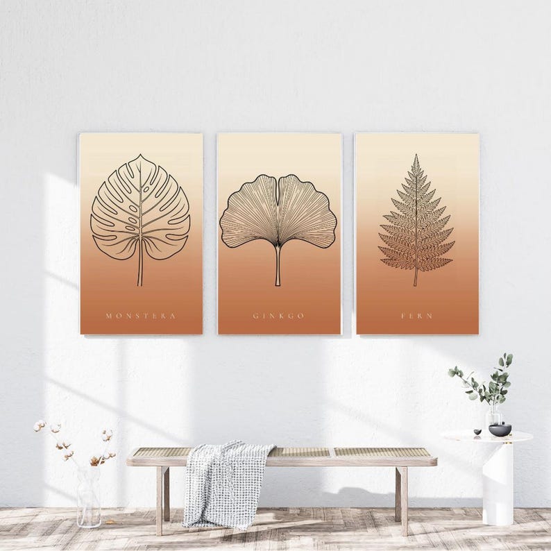 May include: A set of three rectangular wall art prints featuring black line art illustrations of a monstera leaf, a ginkgo leaf, and a fern leaf. Each print has a gradient background transitioning from light cream to a warm terracotta orange.