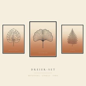 May include: A set of three rectangular wall art prints in black frames, featuring line art of a monstera leaf, a ginkgo leaf, and a fern leaf. Each print has a gradient background transitioning from cream to terracotta. Text includes MONSTERA, GINKGO, FERN, and DREIER-SET.