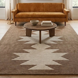 Könnte beinhalten: Ein braun-weißer Teppich mit geometrischem Muster und einem großen, sternartigen Motiv in der Mitte. Der Teppich liegt unter einem runden Couchtisch aus Holz mit einer Schale und Büchern, einem senfgelben Sofa, einer Stehlampe und Wandkunst.