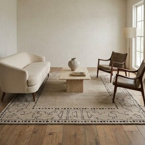 May include: A living room set featuring a cream-colored curved sofa, two dark wood armchairs with gray cushions, a rectangular travertine coffee table with a central vase, and a large area rug with a dark geometric border pattern.