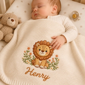 May include: A cream-colored knitted baby blanket featuring an embroidered design of a brown lion surrounded by small orange and white flowers. The name Henry is embroidered in brown cursive script below the lion. A brown teddy bear is nearby.