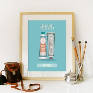 May include: A framed print with a blue background featuring two tubes of toothpaste. The text on the print reads "Give me your best smile".