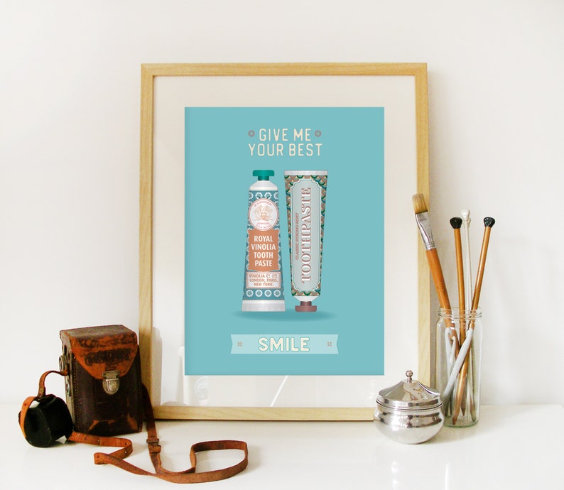 May include: A framed print with a blue background featuring two tubes of toothpaste. The text on the print reads "Give me your best smile".