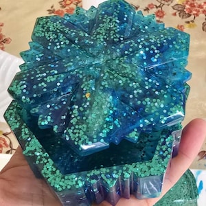 May include: A hexagonal resin trinket box featuring a deep blue and teal colour palette with embedded green glitter. The lid has a geometric, star-like faceted design, and the base has matching angular sides with a glossy, translucent finish.