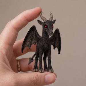 May include: A small, dark charcoal-coloured felted creature with two textured horns, pointed ears, and bright red eyes. The figure features two large, bat-like wings extending from its back and a thin tail ending in a pointed tip, standing on four legs.