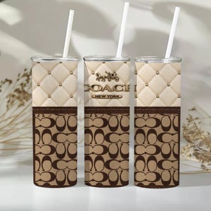 Pode incluir: Três copos cilíndricos com palhinhas brancas, apresentando uma secção superior acolchoada em creme com tachas douradas e uma secção inferior castanha com um padrão repetitivo do logótipo Coach. O copo central exibe o logótipo Coach New York.