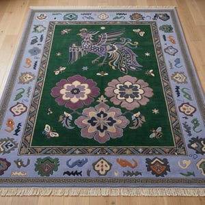 Könnte beinhalten: Ein rechteckiger Teppich mit tiefgrüner Mitte, einem großen violett-blauen Phönix, drei großen violetten Blumenmotiven und kleinen Insekten. Der Rand ist hellblau mit geometrischem Muster, bunten Symbolen und weißen Fransen an den Enden.
