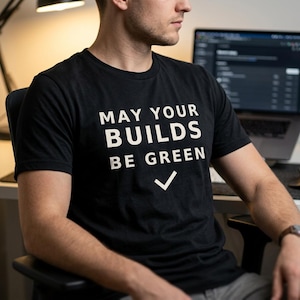 Pode incluir: Uma t-shirt preta de manga curta com o texto branco "MAY YOUR BUILDS BE GREEN" impresso no peito, acompanhado por um símbolo de visto branco posicionado diretamente abaixo do texto, centrado na parte frontal da peça.