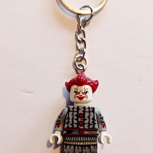 May include: A keyring featuring a small plastic figure of a clown with bright red hair, a white face with red makeup details, and a grey and black patterned outfit. The figure is attached to a silver-coloured metal chain and a circular key ring.