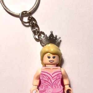 Puede incluir: Un llavero con una minifigura de plástico que lleva un vestido rosa con patrones de remolinos blancos y una corona plateada. La figura tiene cabello rubio, una expresión sonriente y está unida a una anilla metálica por una cadena.