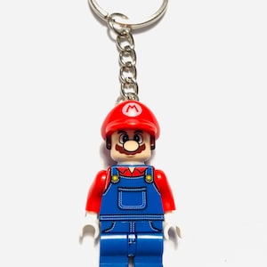 Puede incluir: Un llavero con una figura de plástico de Mario que lleva una gorra roja con un logo M blanco, una camisa roja y un peto azul con botones amarillos. La figura está unida a una anilla de metal plateado mediante una cadena corta de metal.
