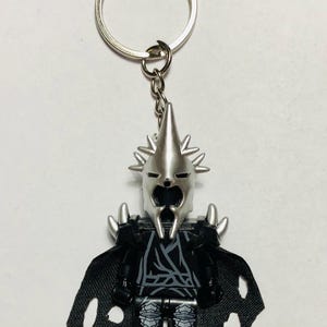 Op de afbeelding: Een zilverkleurige sleutelhanger met een gestileerd figuur met helm en puntige kroon, een zwarte romp met grijze lijndetails, zwarte benen met grijze harnaspatronen en een zwarte stoffen cape met gerafelde randen aan een metalen ring.